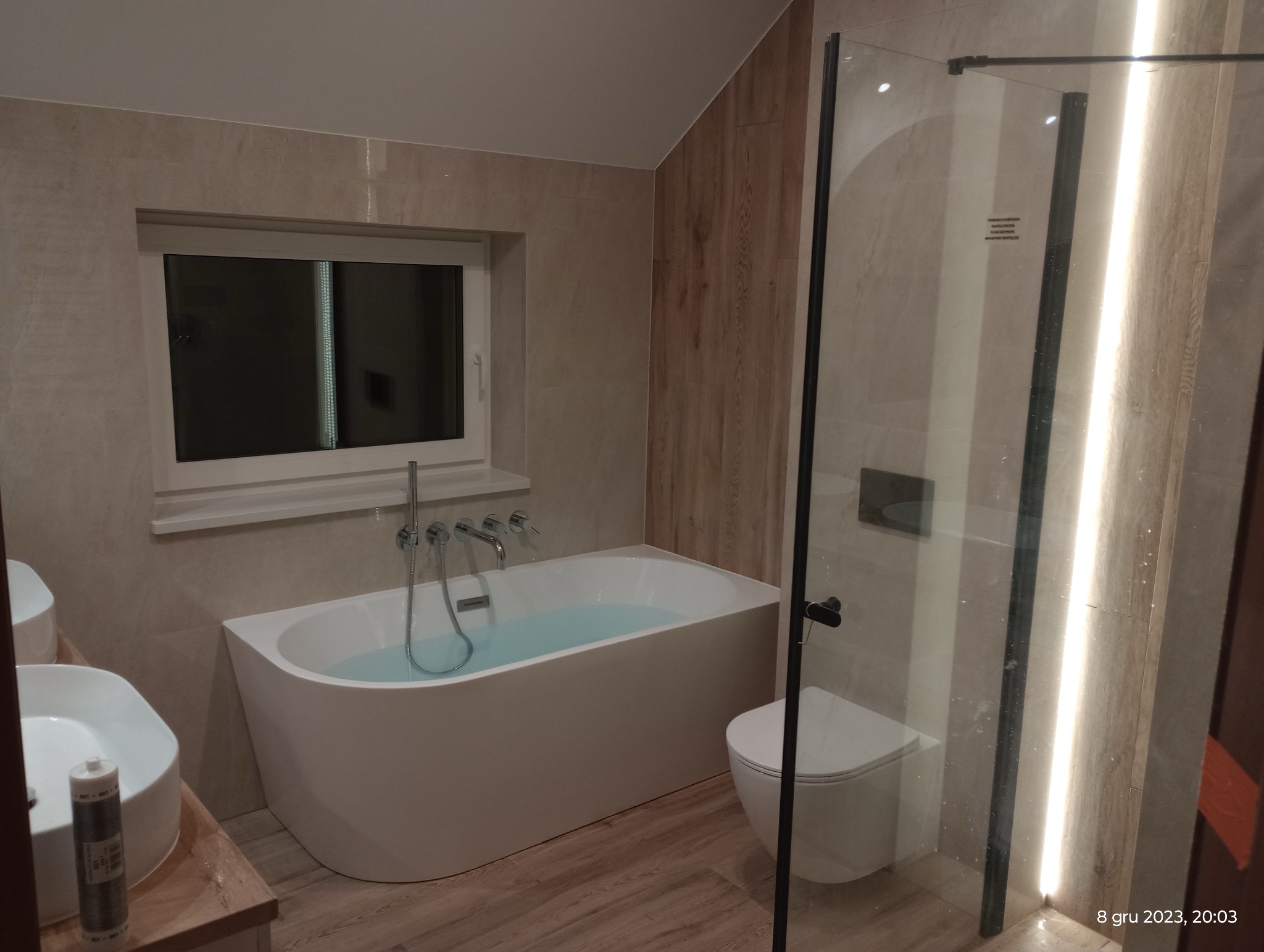 Nowoczesna łazienka z wanną wypełnioną wodą, umywalką na drewnianym blacie i kabiną prysznicową ze szklanymi drzwiami. Beżowe płytki imitujące drewno na ścianach i podłodze.