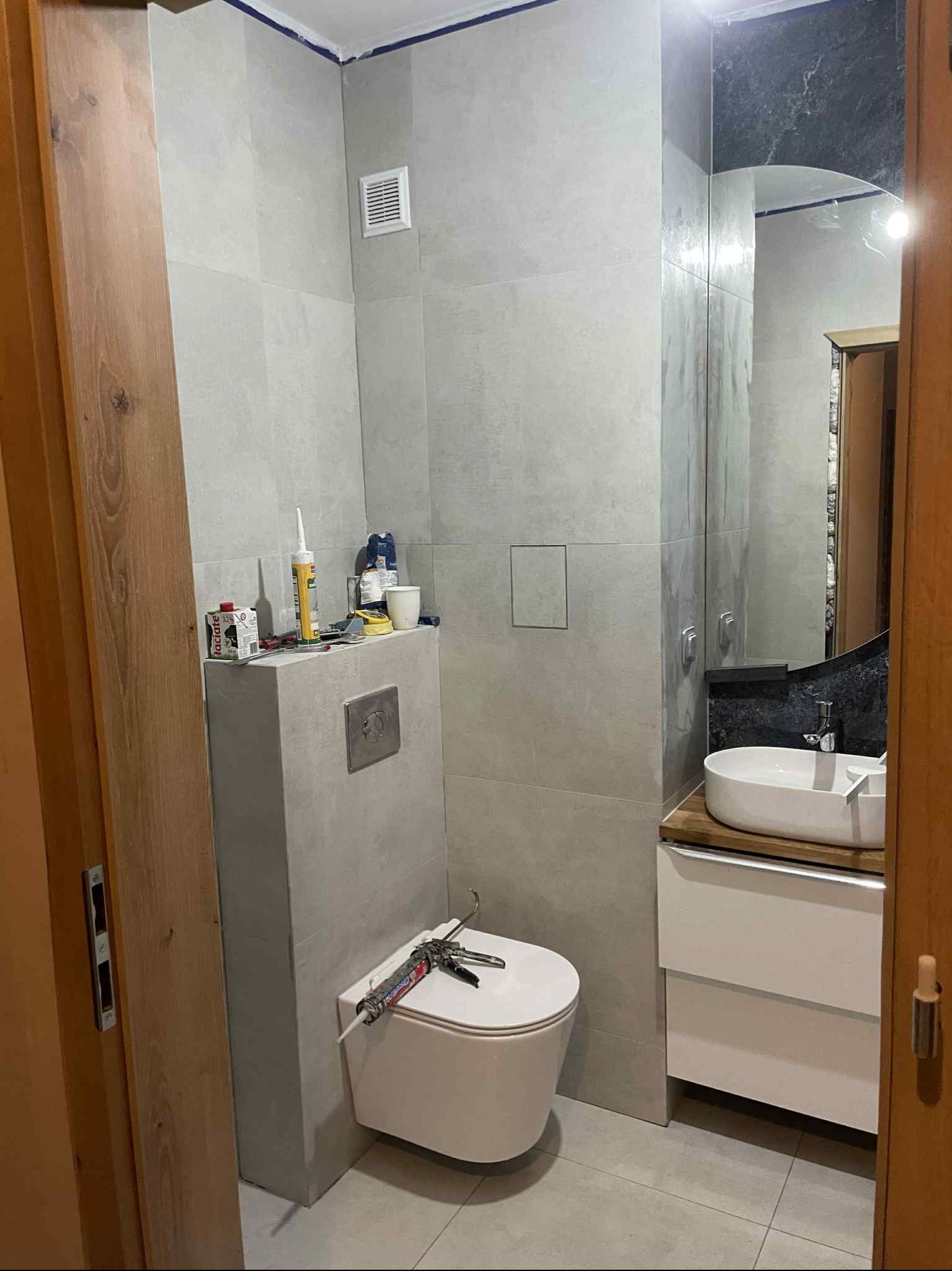 Wykończona łazienka w szarościach z wiszącą toaletą, umywalką na szafce i lustrem. Widoczne narzędzia i materiały budowlane. Minimalistyczny design.