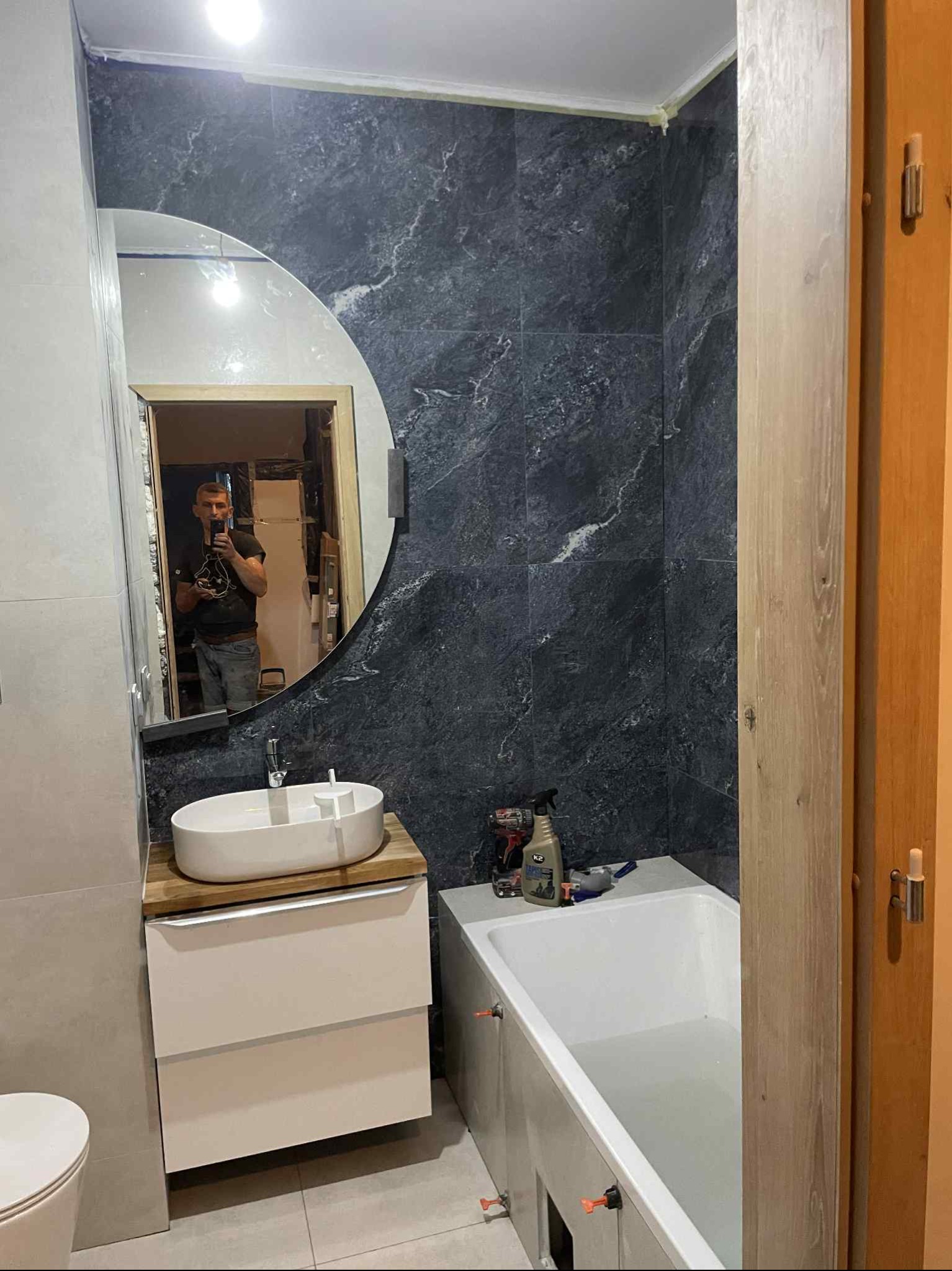 Wykończona łazienka z umywalką na szafce, wanną i ciemnymi płytkami imitującymi marmur na ścianie. W lustrze odbija się osoba robiąca zdjęcie.