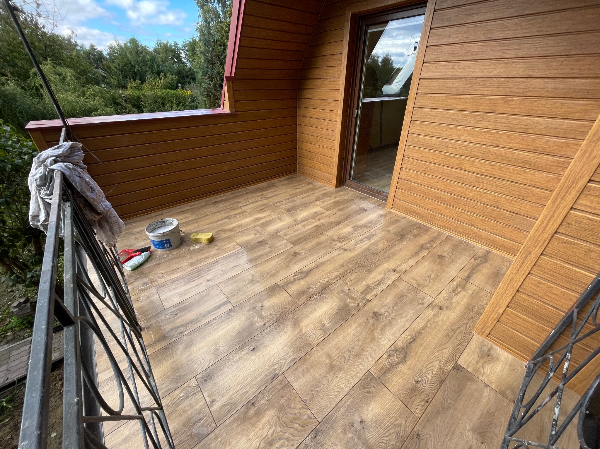 Balkon z drewnopodobną okładziną ścian i podłogi, widoczne narzędzia pracy: wiadro, gąbka. Ujęcie z zewnątrz, zieleń w tle. Realizacja wykończenia balkonu.