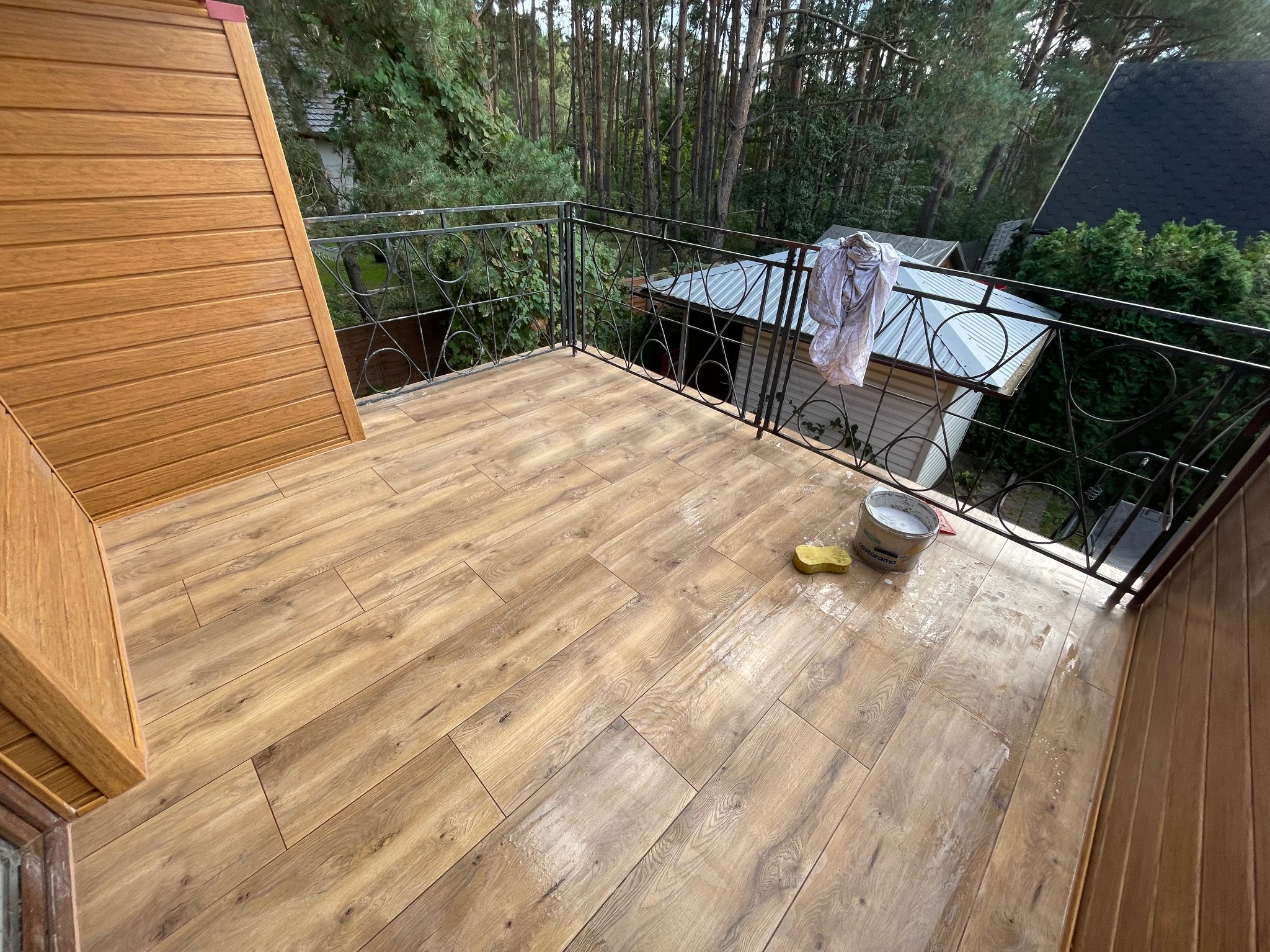 Balkon z drewnopodobną podłogą po czyszczeniu. Widoczne wiadro, gąbka i ślady wilgoci. Metalowa balustrada z dekoracyjnymi elementami. W tle zieleń drzew.