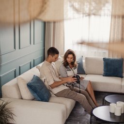 Manufactura - projektowanie wnętrz - Para siedzi na beżowej sofie w salonie z turkusową ścianą. Mężczyzna trzyma aparat, kobieta patrzy na ekran. W tle widać okno z zasłonami i dekoracyjny abażur.