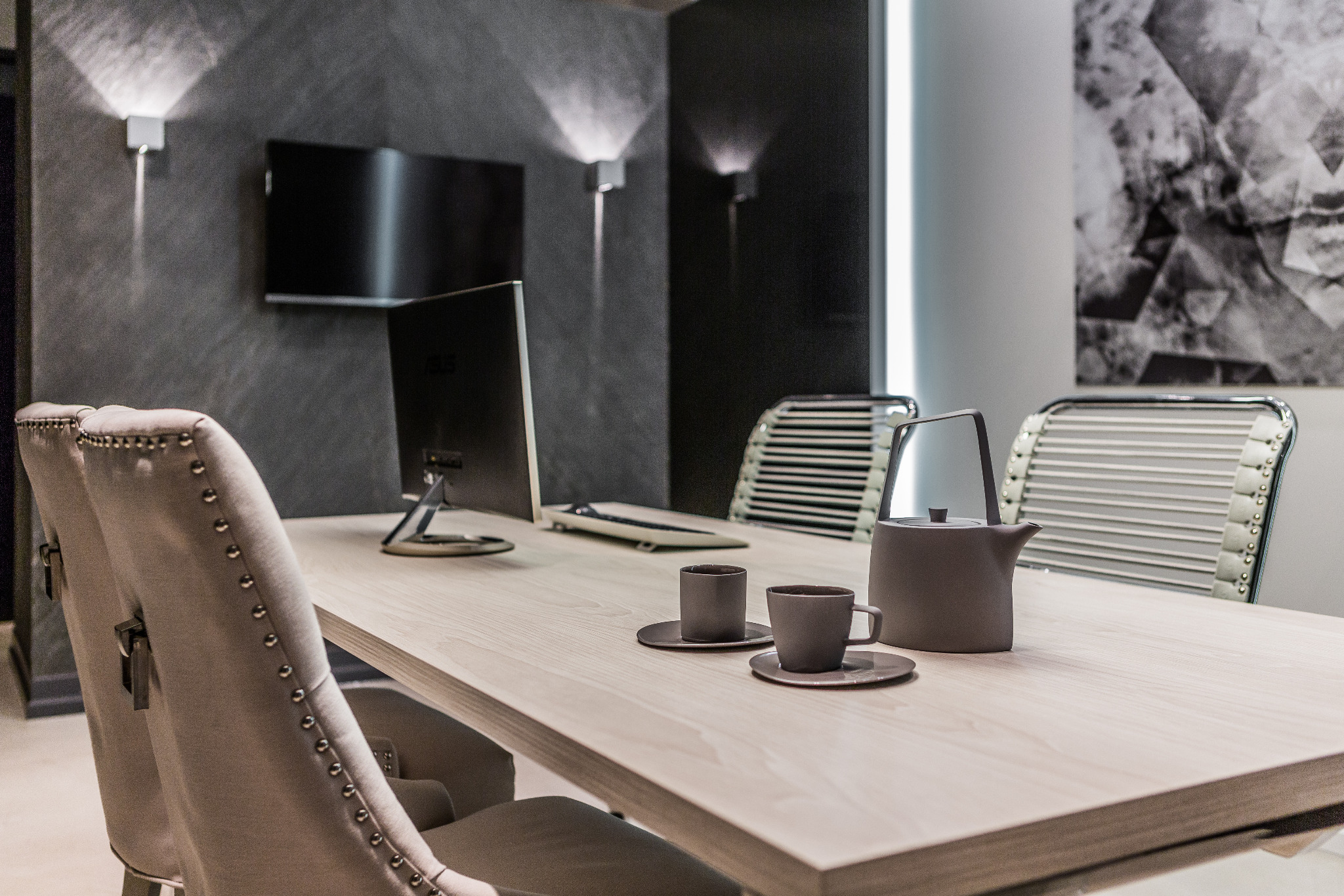 Stół konferencyjny z jasnego drewna z szarym serwisem do kawy, krzesła z tapicerką w stylu glamour, monitor i lampy ścienne w nowoczesnym biurze.