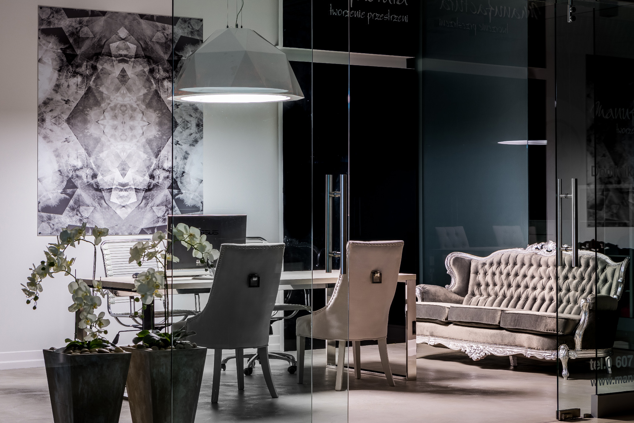 Nowoczesne biuro z szklaną ścianą, designerską lampą, biurkiem, krzesłami i sofą w stylu glamour, uzupełnione abstrakcyjnym obrazem i storczykami w donicach.