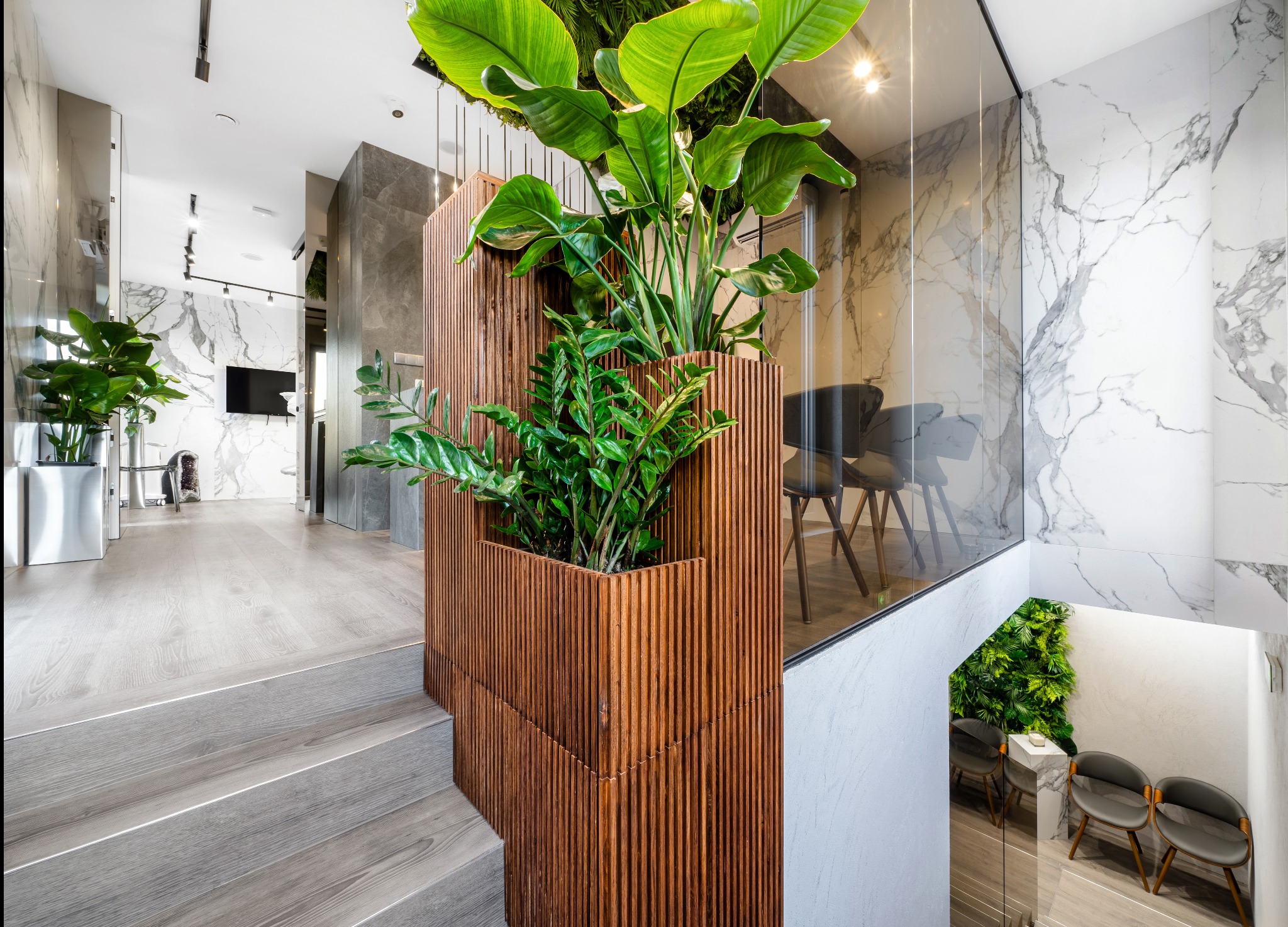 Nowoczesne wnętrze z drewnianą ścianką działową, roślinami i marmurowymi wykończeniami. Widok z góry na schody i przestrzeń biurową z krzesłami.
