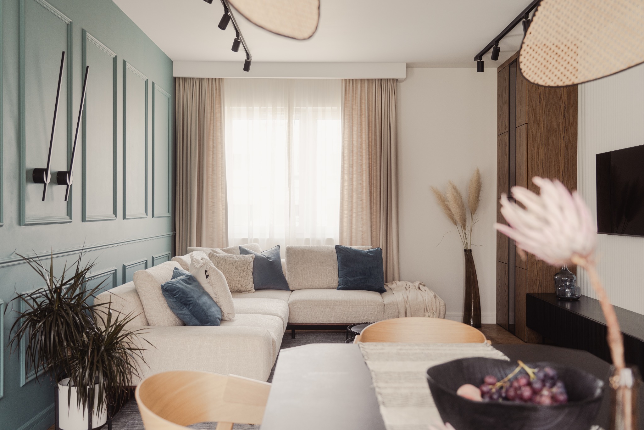 Jasny salon z narożnikiem, niebieskimi poduszkami i dekoracyjną ścianą w kolorze miętowym. Na stole misa z owocami, w tle okno z beżowymi zasłonami i lampa sufitowa.