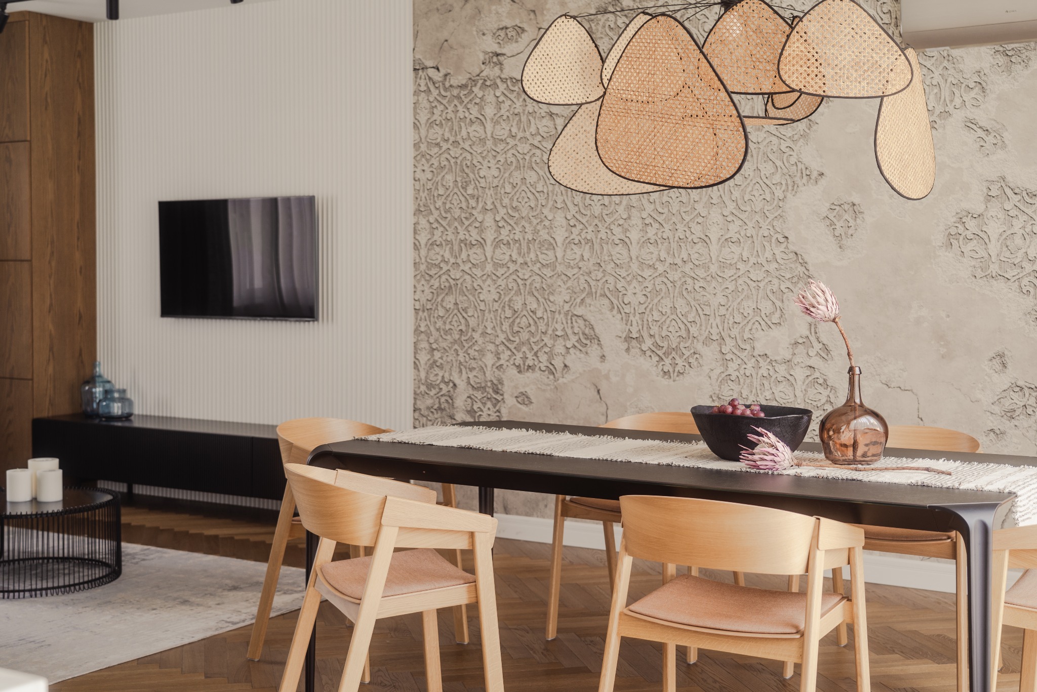 Stylowa jadalnia z drewnianymi krzesłami, czarnym stołem i dekoracyjną lampą z rattanu. Na stole misa z owocami i wazon z kwiatem. W tle ściana z dekoracyjnym wzorem.