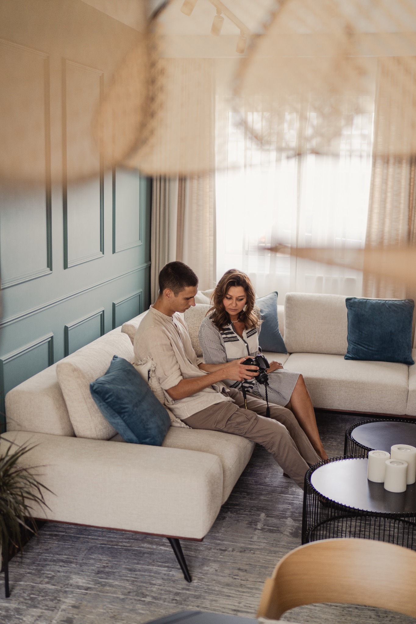 Para siedzi na beżowej sofie w salonie z turkusową ścianą. Mężczyzna trzyma aparat, kobieta patrzy na ekran. W tle widać okno z zasłonami i dekoracyjny abażur.