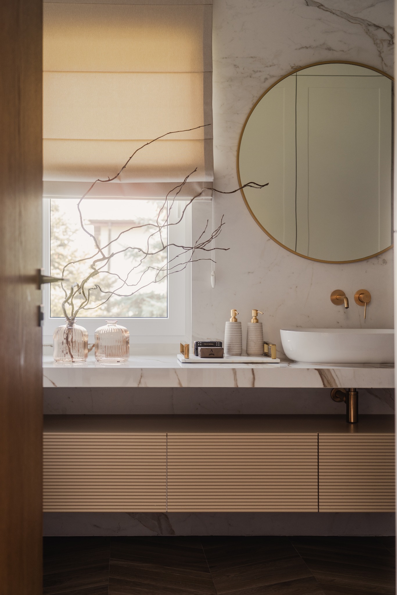 Elegancka łazienka z marmurową umywalką, okrągłym lustrem w złotej ramie i beżowymi roletami rzymskimi. Dekoracyjne gałązki w wazonach dodają naturalnego akcentu.