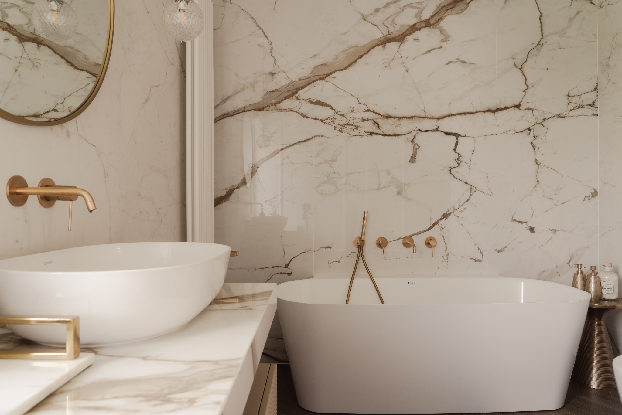 Elegancka łazienka z marmurowymi płytkami, owalną umywalką i wanną. Złote baterie i dodatki podkreślają luksusowy charakter wnętrza. Minimalistyczny design z dbałością o detale.