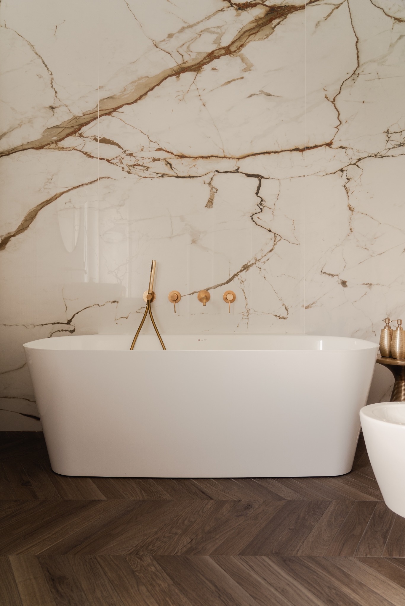 Elegancka łazienka z wolnostojącą wanną na tle marmurowej ściany i parkietu ułożonego w jodełkę. Złota armatura i dodatki podkreślają luksusowy charakter.