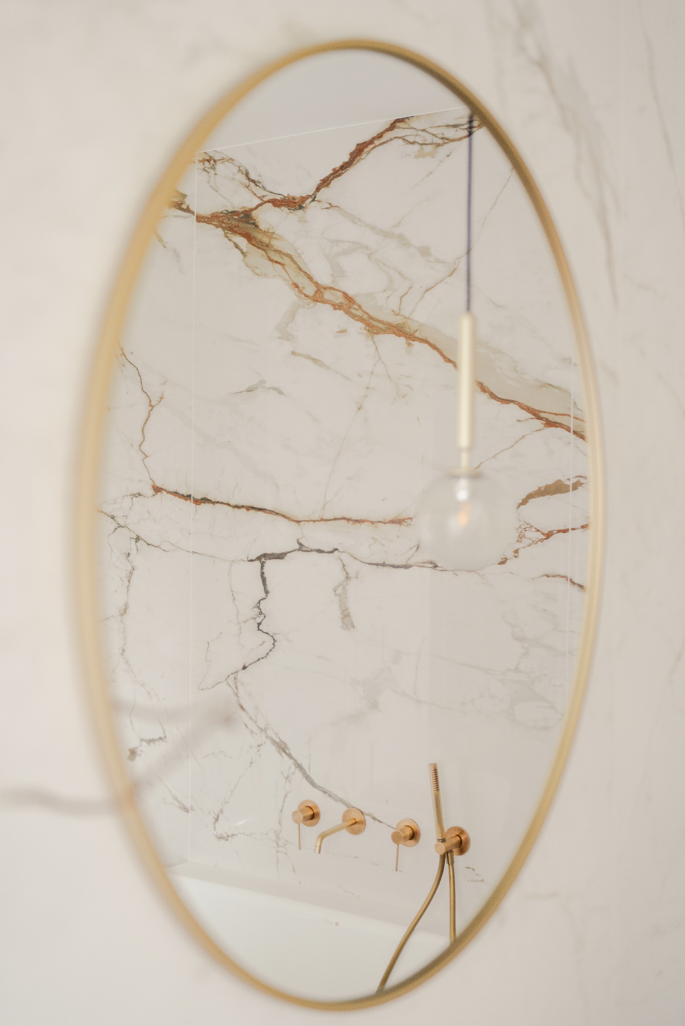 Owalne lustro z cienką, złotą ramą odbija fragment łazienki: białe marmurowe płytki, złotą armaturę i wiszącą lampę z okrągłym kloszem. Minimalistyczny design.