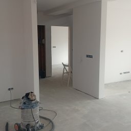 Set-Bud usługi remontowo-wykończeniowe - Wnętrze w trakcie prac wykończeniowych: białe ściany, surowa podłoga, widoczny odkurzacz przemysłowy i drabina. Przygotowanie do dalszych etapów remontu.