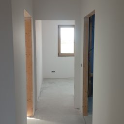 Set-Bud usługi remontowo-wykończeniowe - Wnętrze w trakcie wykańczania: białe ściany, surowa podłoga, widoczne ościeżnice drzwiowe z surowego drewna, okno z drewnianą ramą w głębi pomieszczenia.