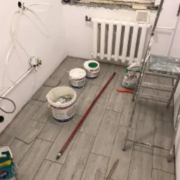 Remont łazienki w trakcie: układanie jasnoszarych płytek imitujących drewno, widoczne wiaderka z farbą, drabina malarska, grzejnik, puszki elektryczne z kablami oraz okno z parapetem w stanie surowym.