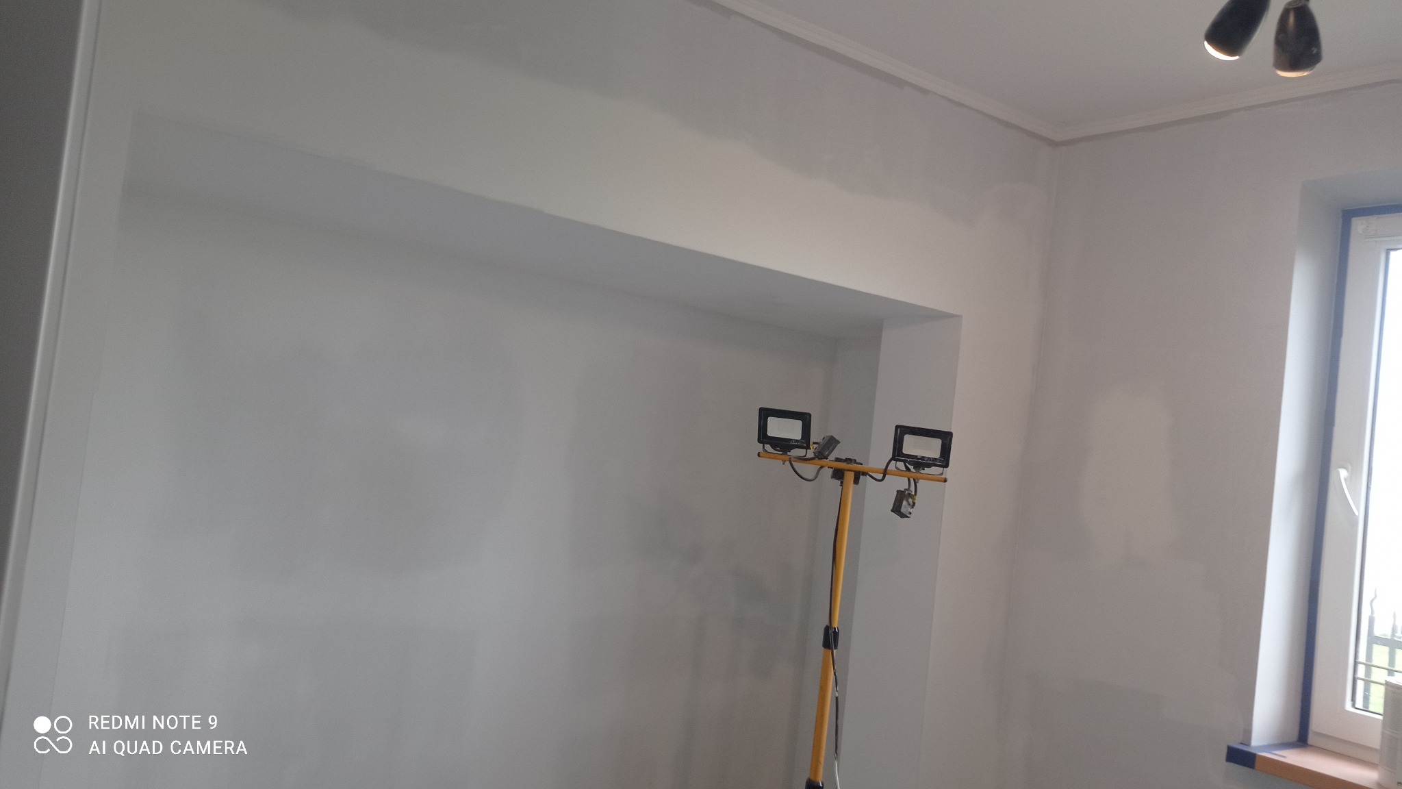 Świeżo zagruntowane ściany w pokoju z wnęką, widoczne ślady po szpachlowaniu, na podłodze lampa budowlana, okno z niebieskimi akcentami.
