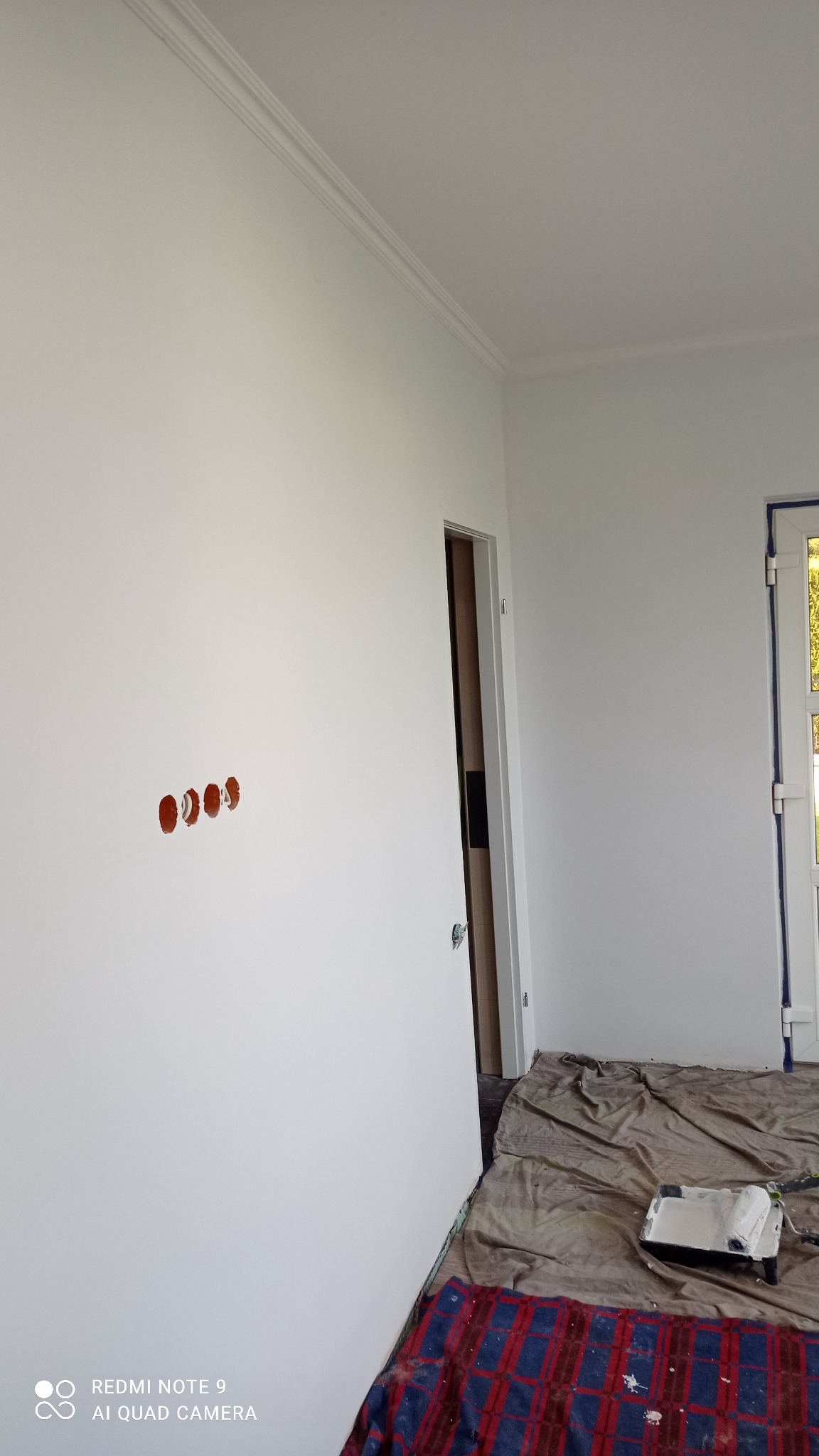 Świeżo pomalowane na biało ściany z listwą ozdobną pod sufitem, widoczne puszki elektryczne przygotowane do montażu osprzętu oraz zabezpieczona folią podłoga z wałkiem i kuwetą malarską.