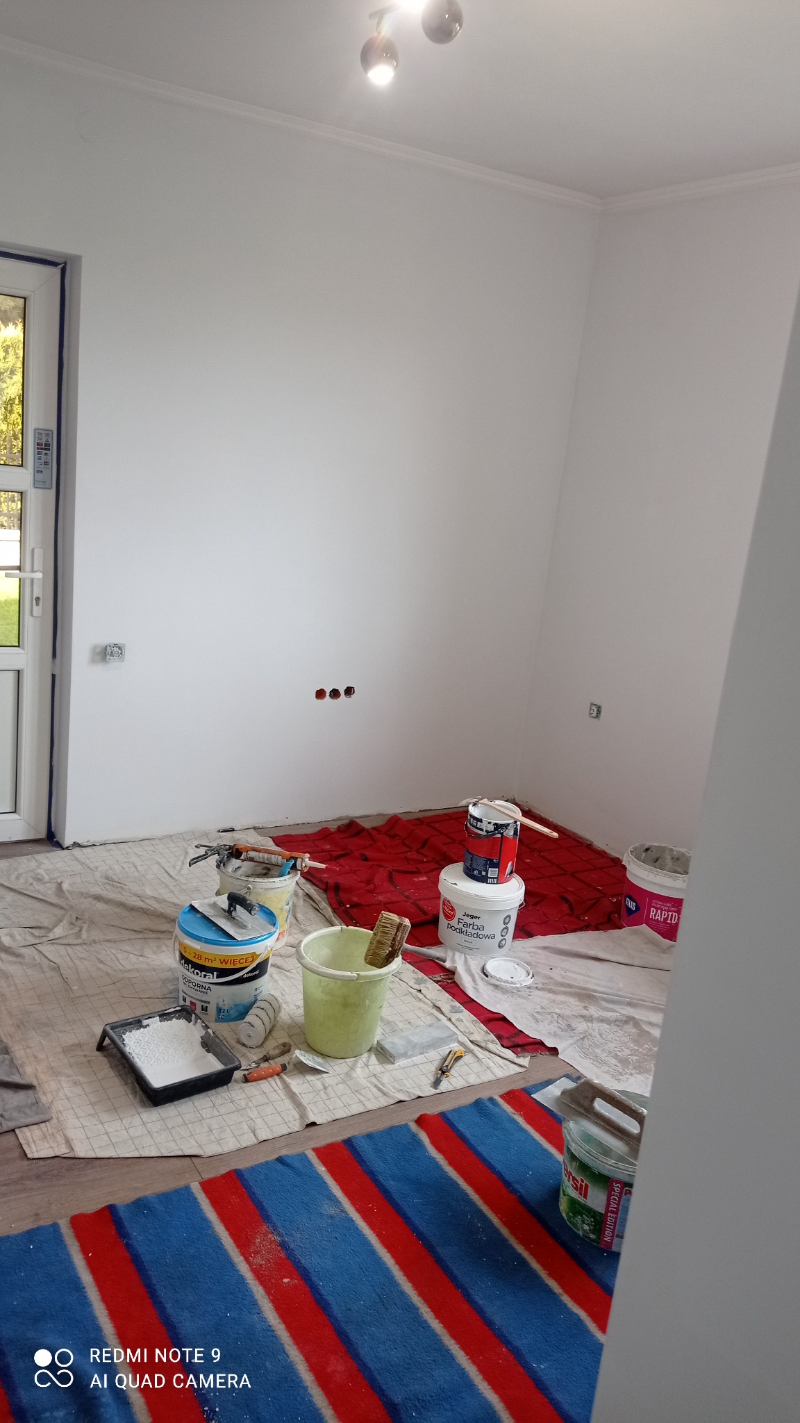 Pokój w trakcie odświeżania, z białymi ścianami, zabezpieczoną podłogą, wiadrami z farbą, pędzlem, wałkiem i kuwetą na podłodze, widoczne puszki farb Dekoral, Jeger i Atlas Rapid.