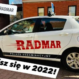 Biały samochód egzaminacyjny OSK Radmar z napisem 'Zapisz się w 2022!' na tle ceglanego budynku.