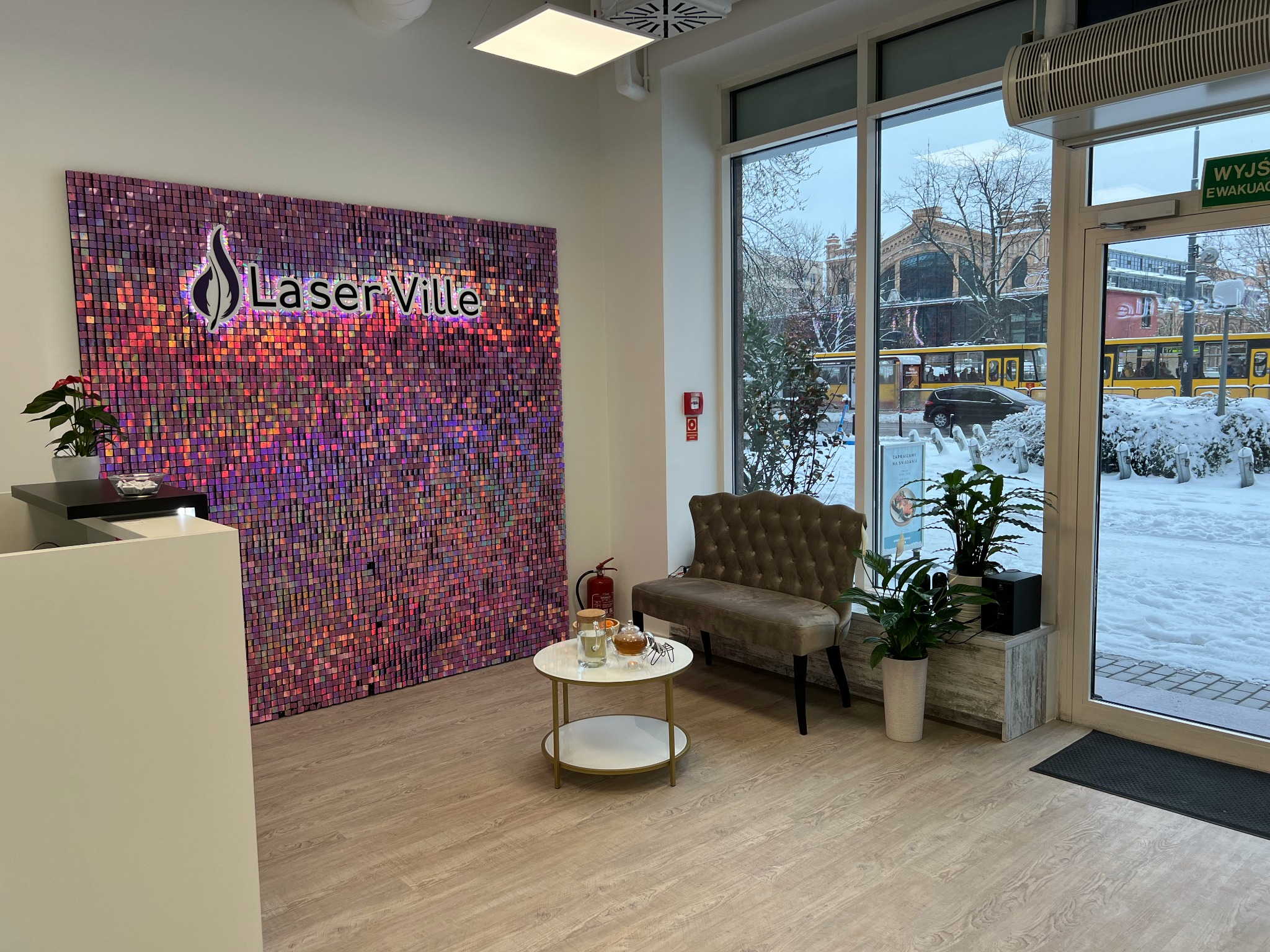 Recepcja salonu kosmetycznego Laser Ville z dekoracyjną ścianą z połyskujących, fioletowo-różowych kwadratów, stolikiem z napojami i widokiem na zaśnieżony krajobraz miejski przez okno.