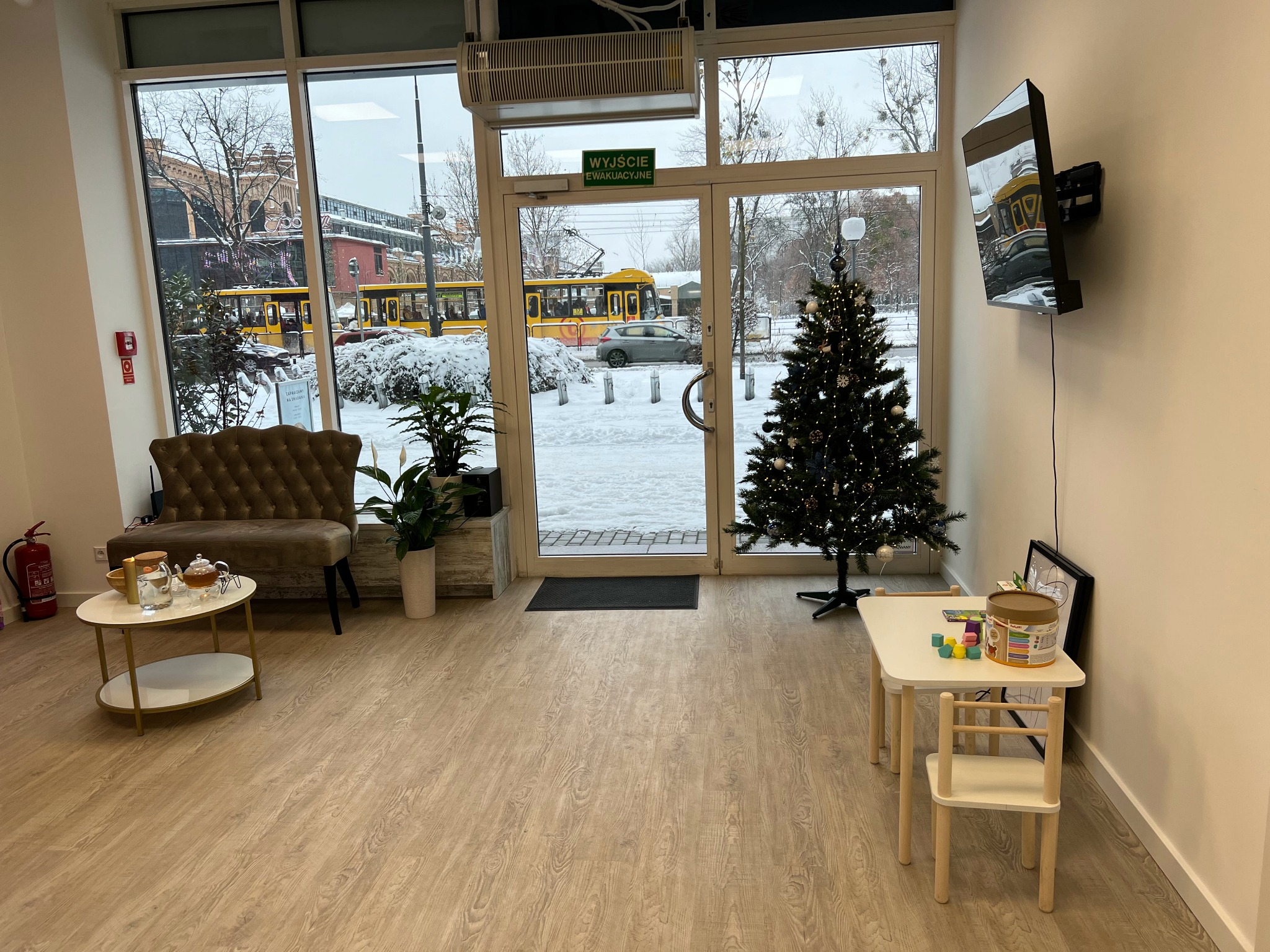 Wnętrze poczekalni z widokiem na zaśnieżony krajobraz miejski, żółty tramwaj za oknem, kanapa, stolik z herbatą i choinka.