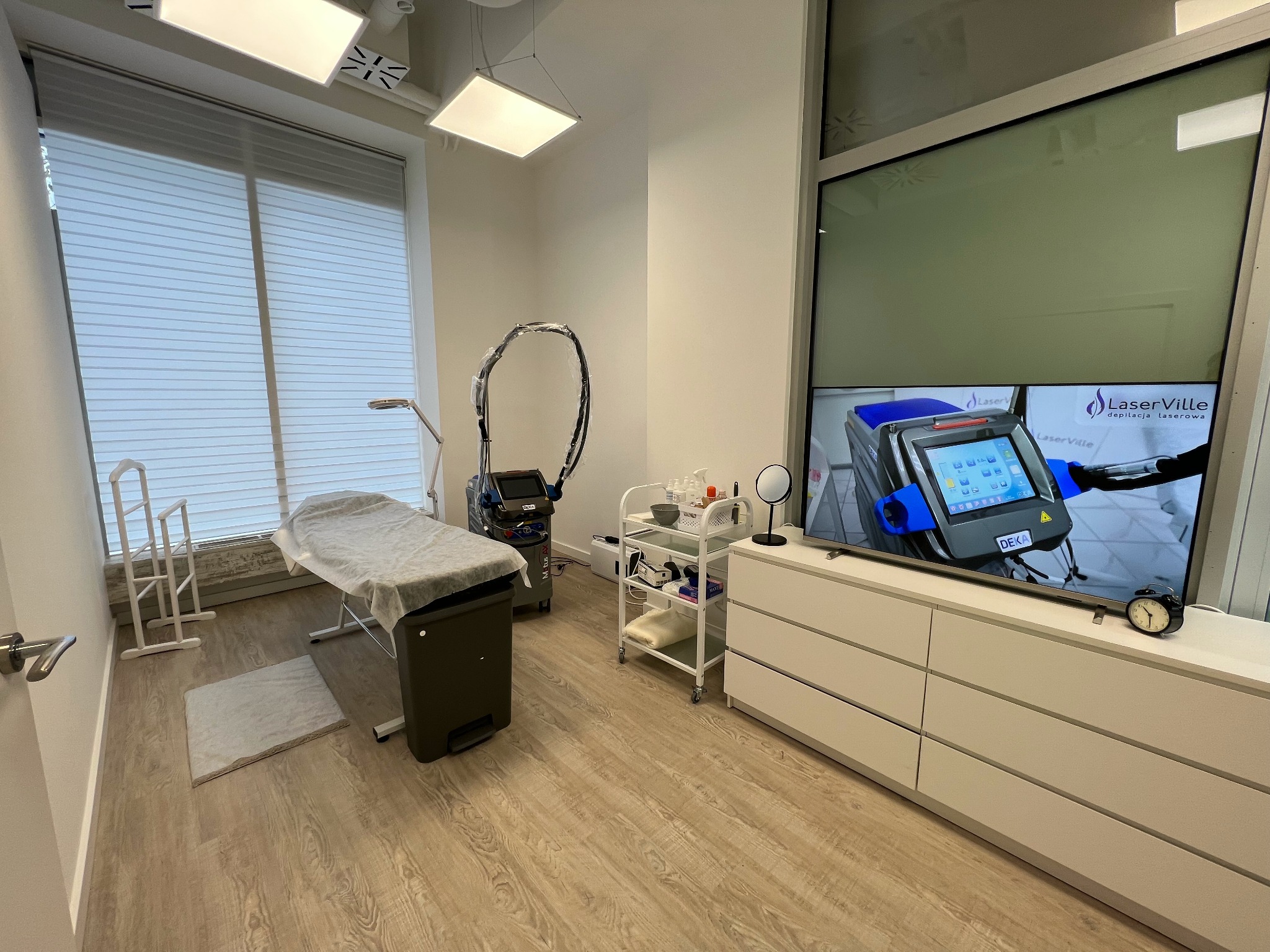 Jasne wnętrze gabinetu kosmetycznego z łóżkiem do zabiegów przykrytym prześcieradłem, urządzeniem do depilacji laserowej i włączonym telewizorem prezentującym urządzenie LaserVille do depilacji...