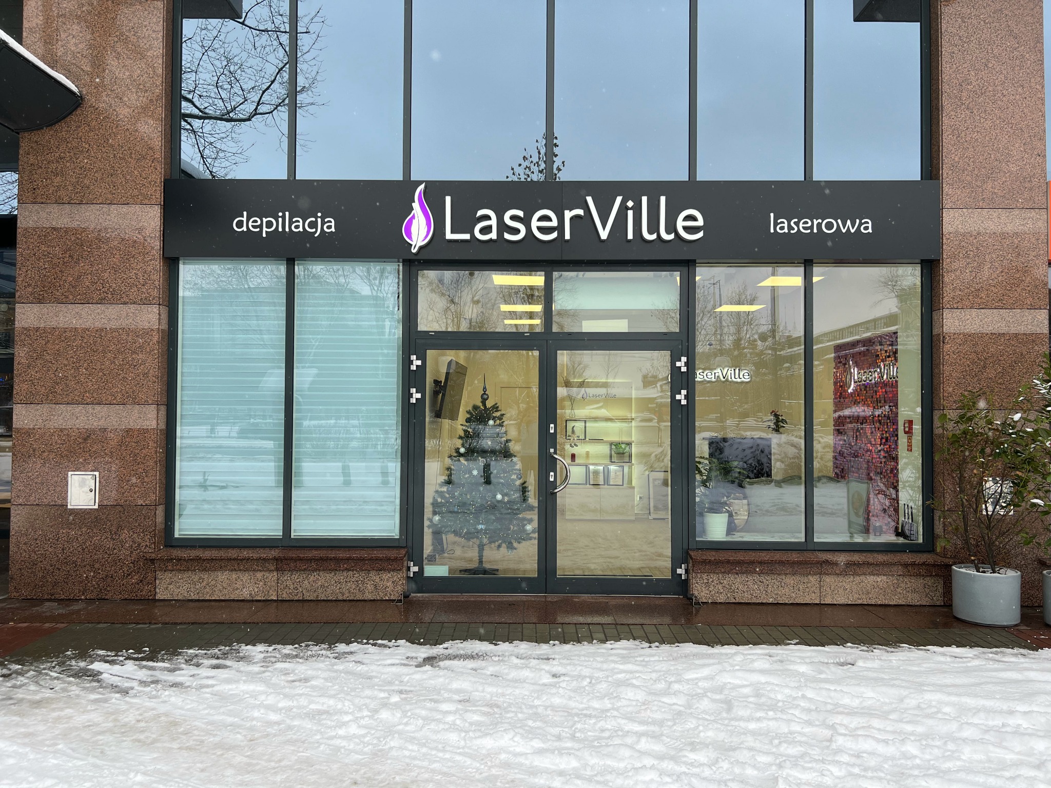 Fasada budynku z napisem LaserVille, oferującego depilację laserową; widoczna choinka wewnątrz lokalu i śnieg na chodniku.