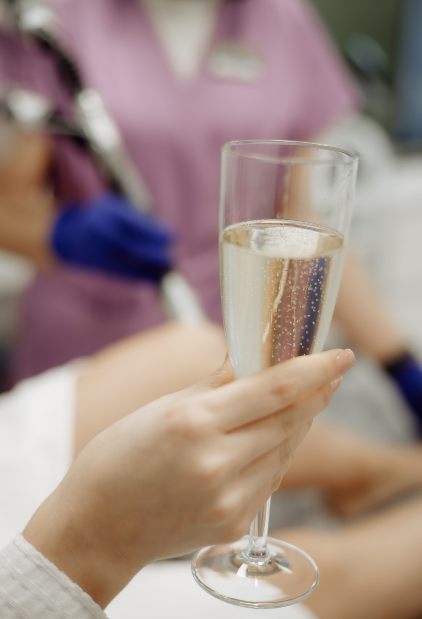 Dłoń trzyma kieliszek z musującym napojem, w tle osoba w uniformie kosmetycznym i niebieskich rękawiczkach, prawdopodobnie podczas relaksującego zabiegu.
