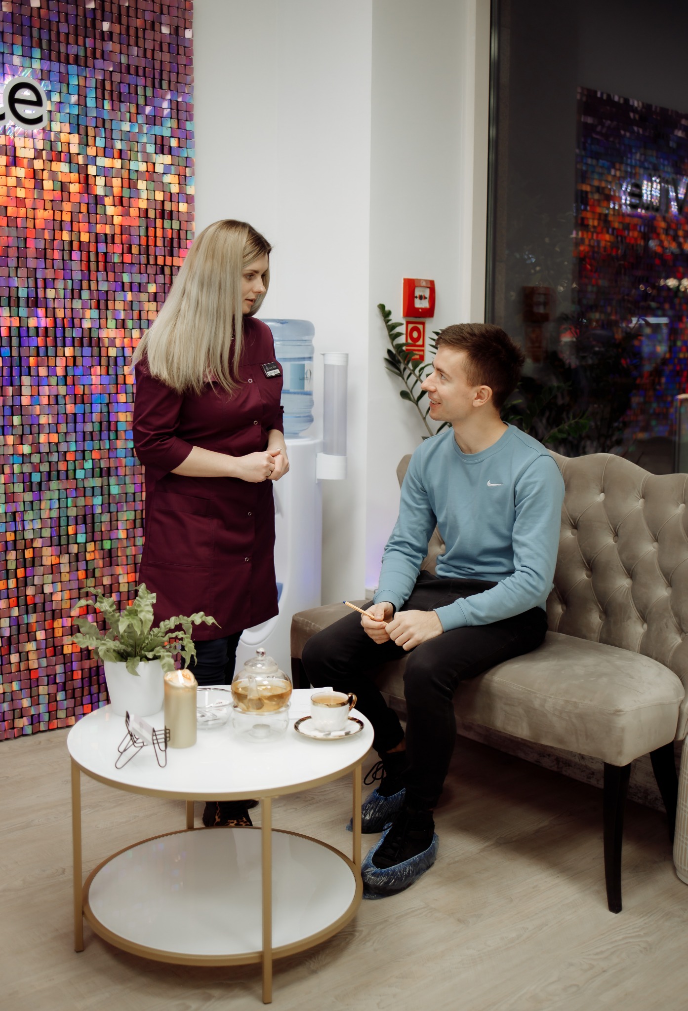 Klient w niebieskich ochraniaczach na obuwiu siedzi na welurowej kanapie w gabinecie, rozmawiając z kosmetyczką w bordowym uniformie, na stoliku herbata i dekoracje.