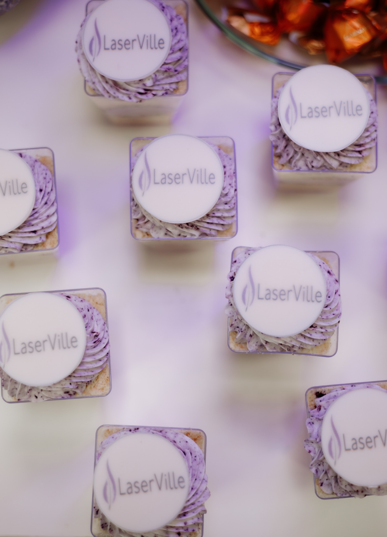 Deserki w przezroczystych pojemnikach z kremem jagodowym i białym okrągłym topperem z logo LaserVille, ułożone na białym stole, w tle naczynie z pomarańczowymi słodkościami.