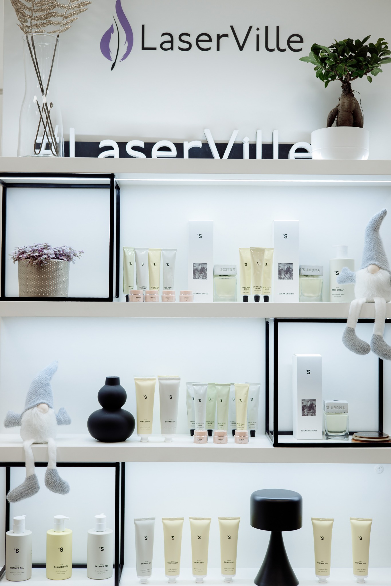 Półki w salonie kosmetycznym z białymi kosmetykami marki 'S' w tubach i butelkach, dekoracjami roślinnymi i figurką skrzata w szarym ubranku.
