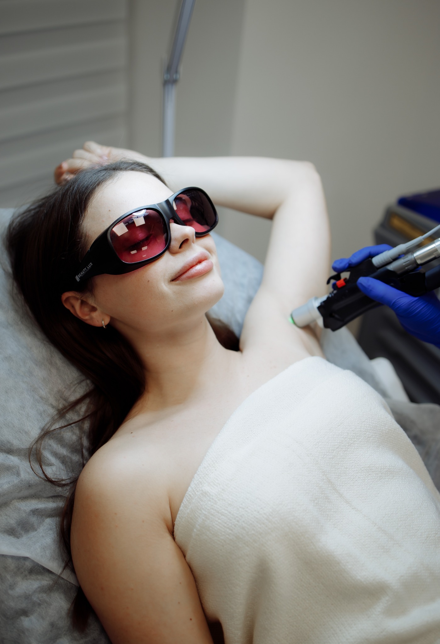 Kobieta w okularach ochronnych poddawana zabiegowi depilacji laserowej pod pachami, widoczny aplikator lasera w dłoni w niebieskiej rękawiczce.