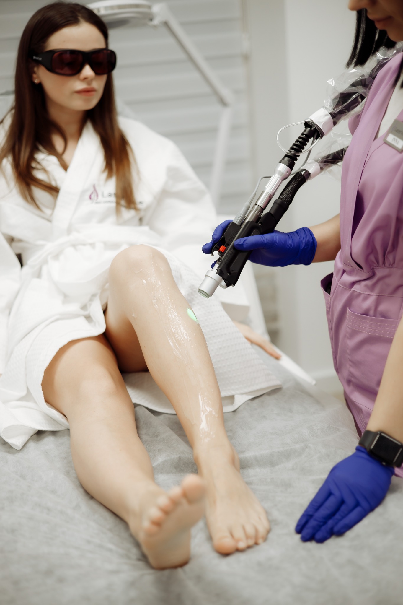 Laserowe usuwanie owłosienia z nogi pacjentki w okularach ochronnych, aplikacja żelu chłodzącego, widoczny zielony promień lasera.