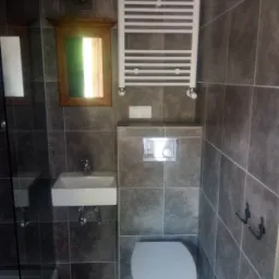 Szara łazienka z kwadratowymi płytkami, umywalką nablatową, toaletą podwieszaną, grzejnikiem drabinkowym i lustrem w drewnianej ramie.