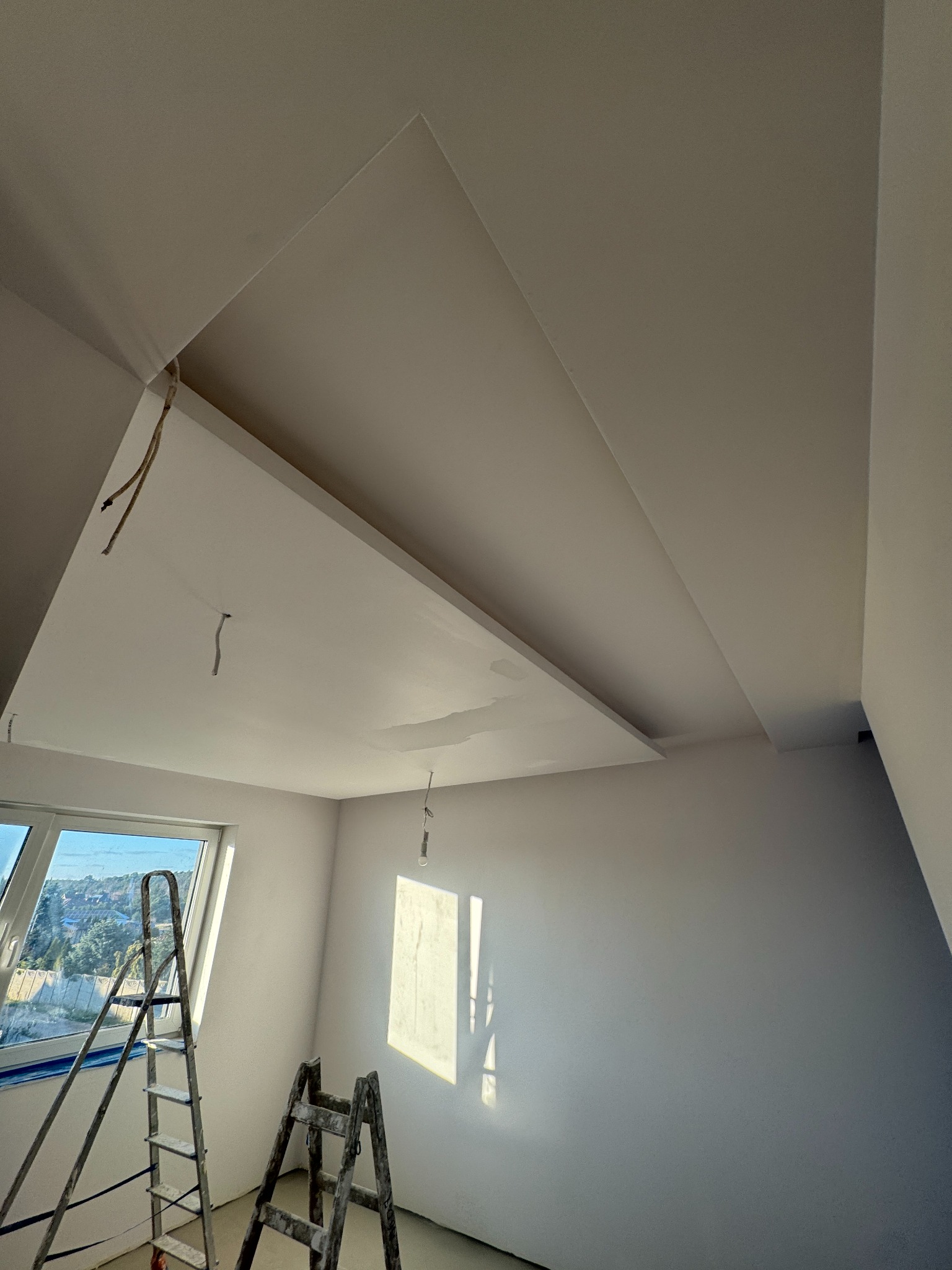 Białe wnętrze w trakcie wykańczania: sufit z płyt karton-gips, drabiny malarskie przy oknie. Widoczne kable i niedoskonałości przed ostatecznym szpachlowaniem.