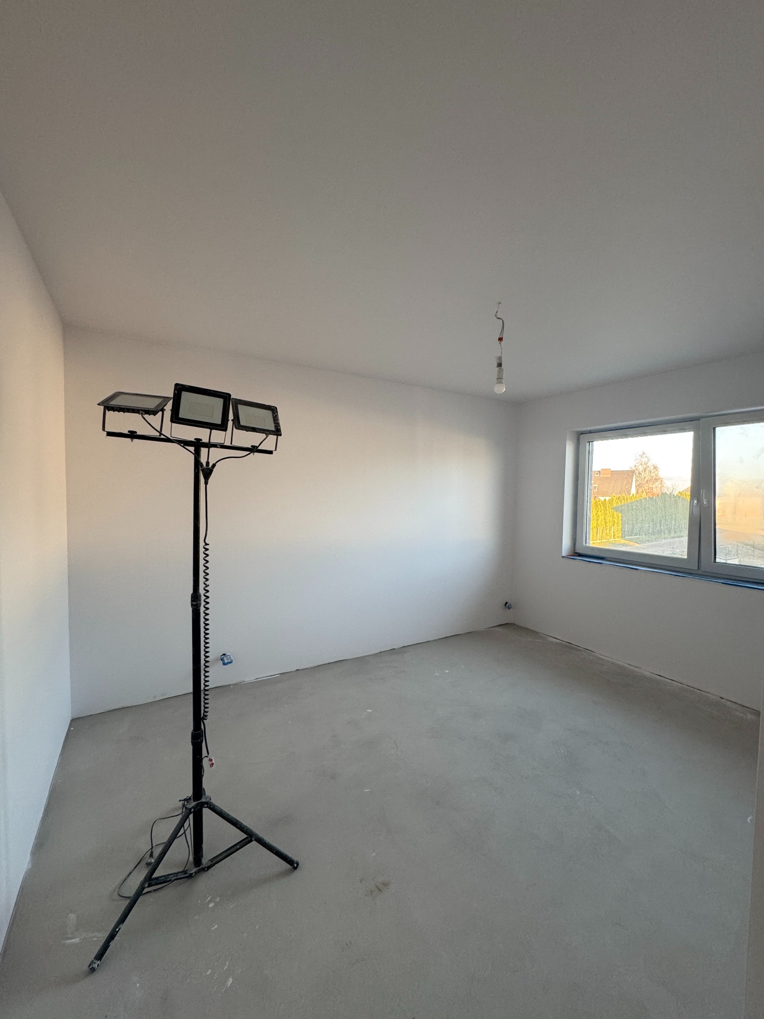 Wnętrze pokoju po wylewce, z widocznym oświetleniem budowlanym na statywie. Ściany przygotowane do dalszych prac wykończeniowych, okno z widokiem na zewnątrz.