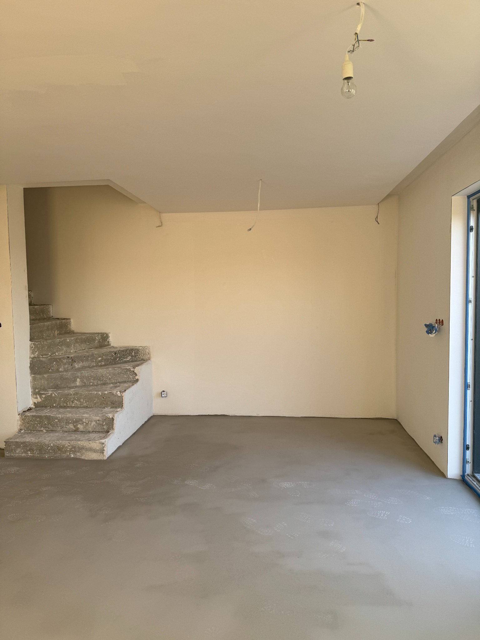 Wnętrze w trakcie prac wykończeniowych: surowe schody, świeżo wylana posadzka z widocznymi odciskami butów, ściany przygotowane do malowania, wisząca żarówka.