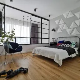 Nowoczesna sypialnia z drewnianą podłogą, czarną ramą okienną, geometryczną dekoracją ścienną i narzędziami do montażu widocznymi na pierwszym planie.