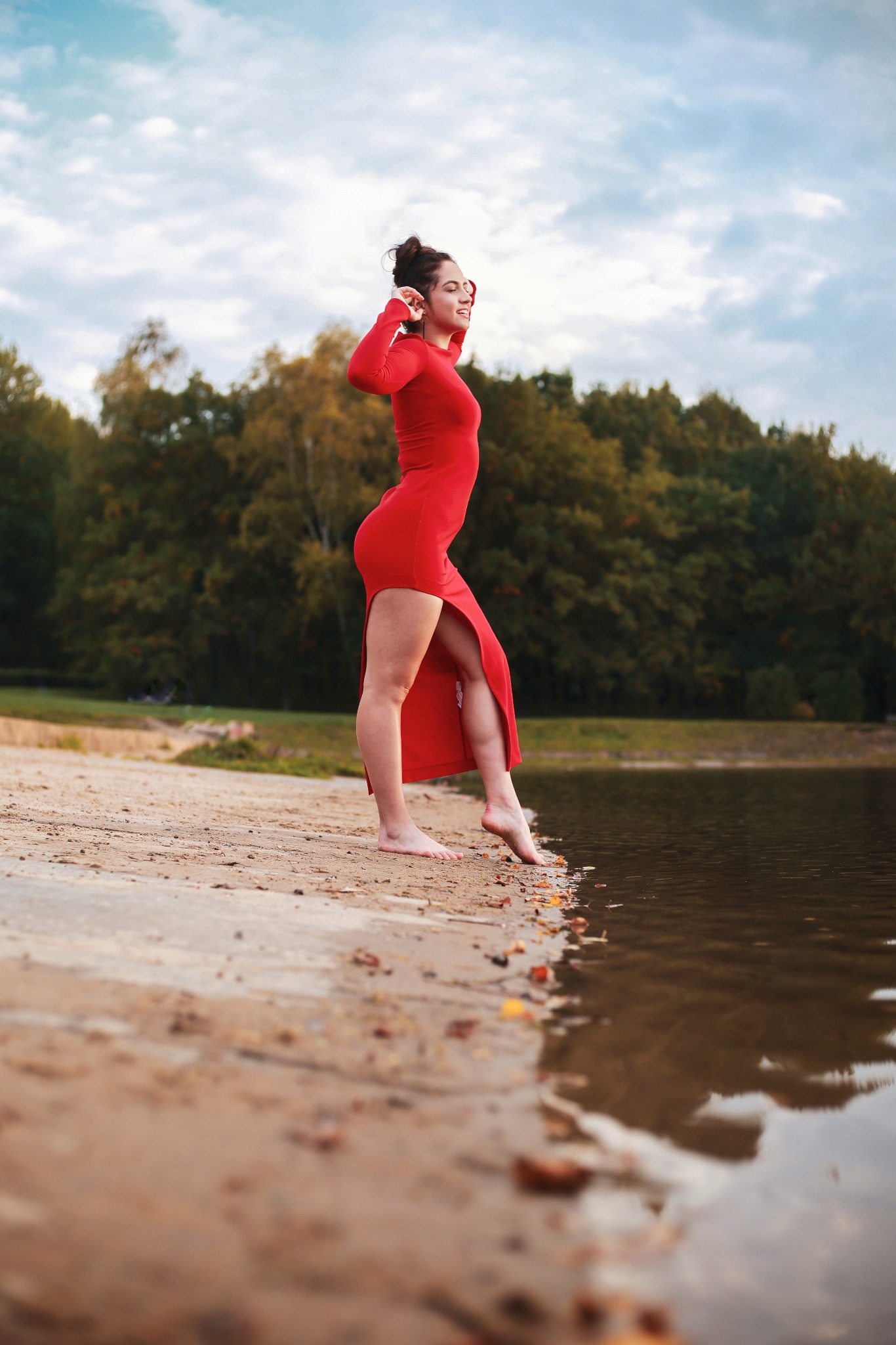 Kobieta w czerwonej sukience z rozcięciem, stojąca boso na brzegu jeziora, z lasem w tle i pochmurnym niebem.