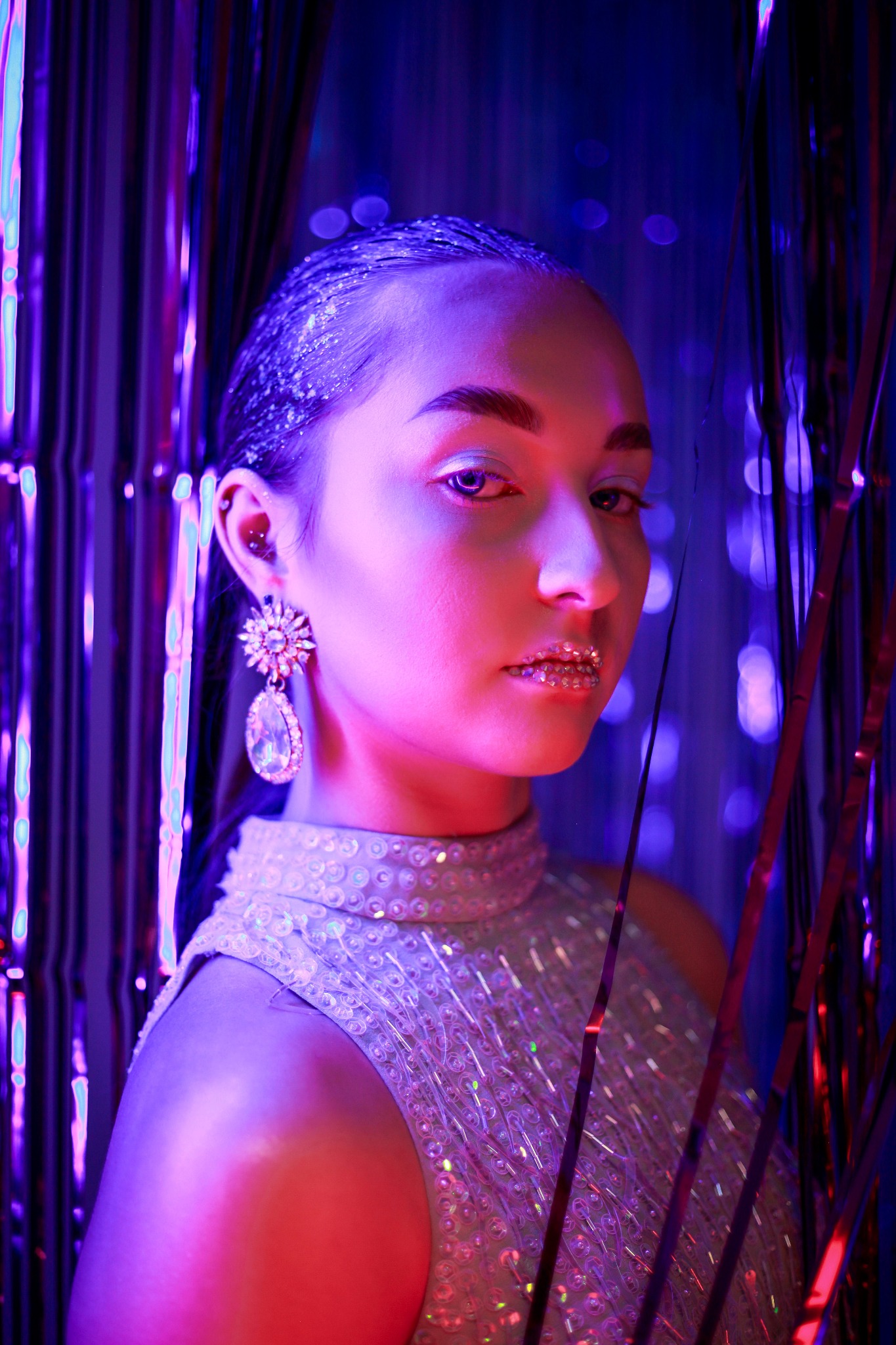 Portret kobiety z brokatem na ustach i włosach, ubrana w cekinową sukienkę na tle błyszczącej kurtyny w neonowym oświetleniu.