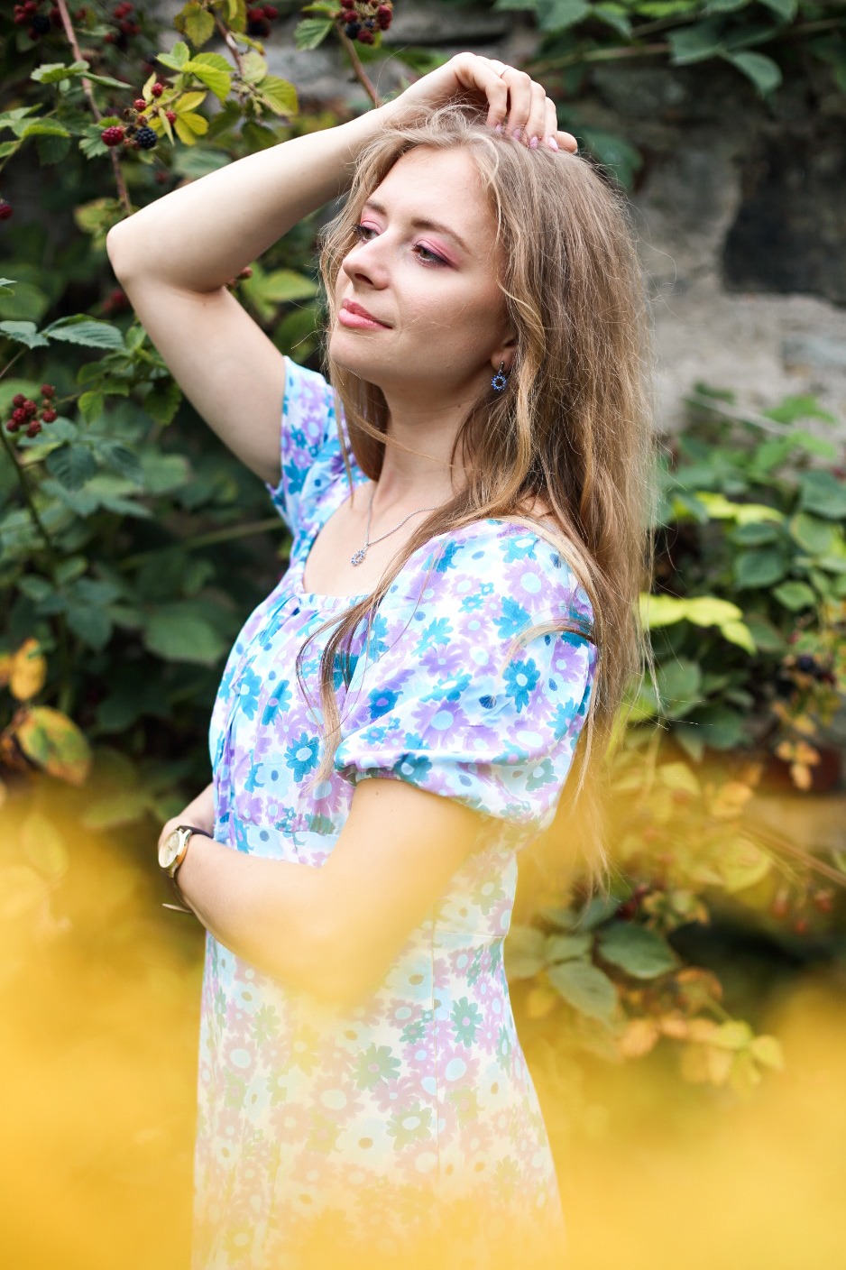 Kobieta w kwiecistej sukience pozuje na tle zieleni i kamiennego muru, z ręką uniesioną do włosów i delikatnym uśmiechem, w miękkim, naturalnym świetle.