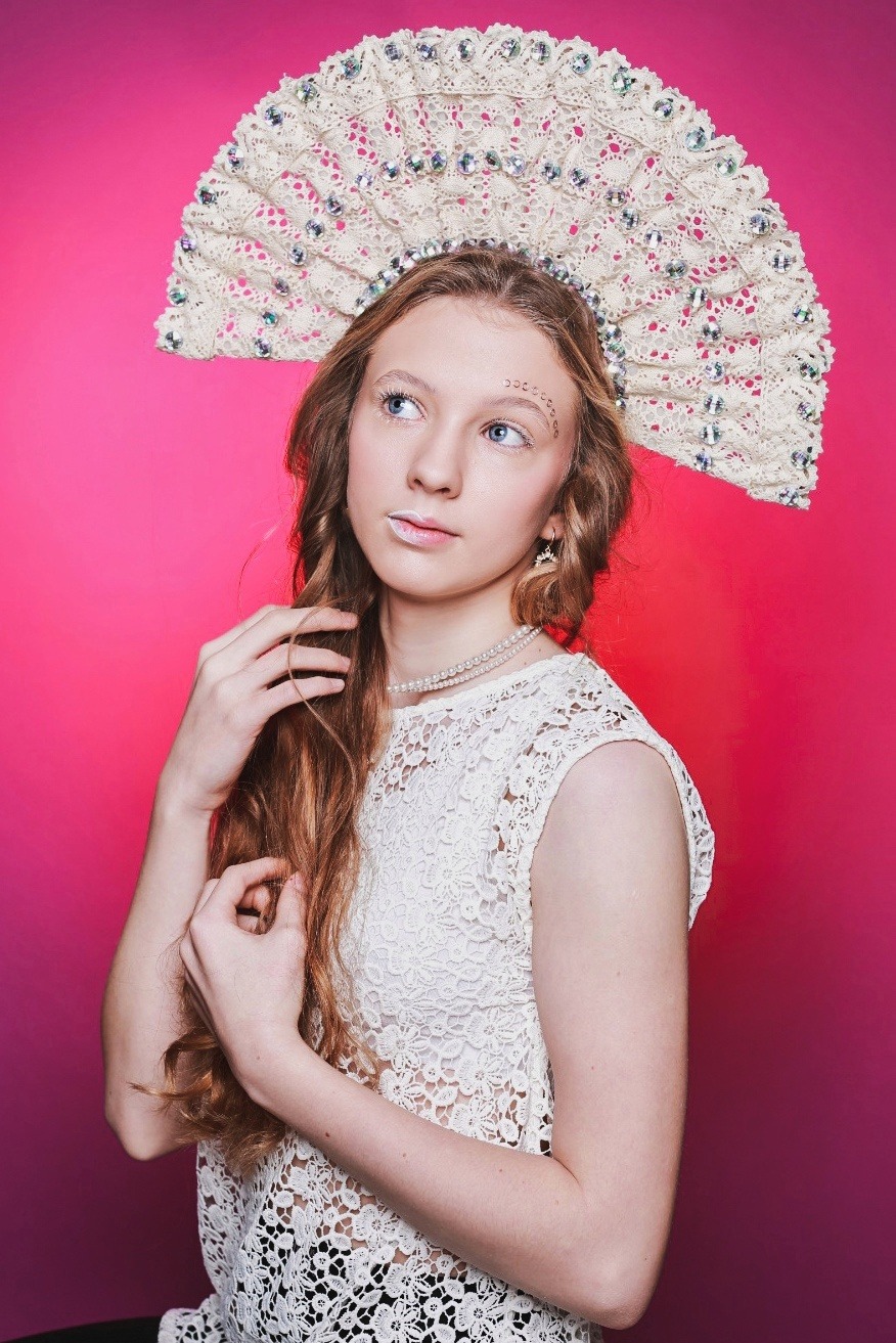 Portret młodej kobiety w koronkowej sukni i bogatym, koronkowym nakryciu głowy na tle różowego gradientu. Delikatny makijaż i perłowy naszyjnik.