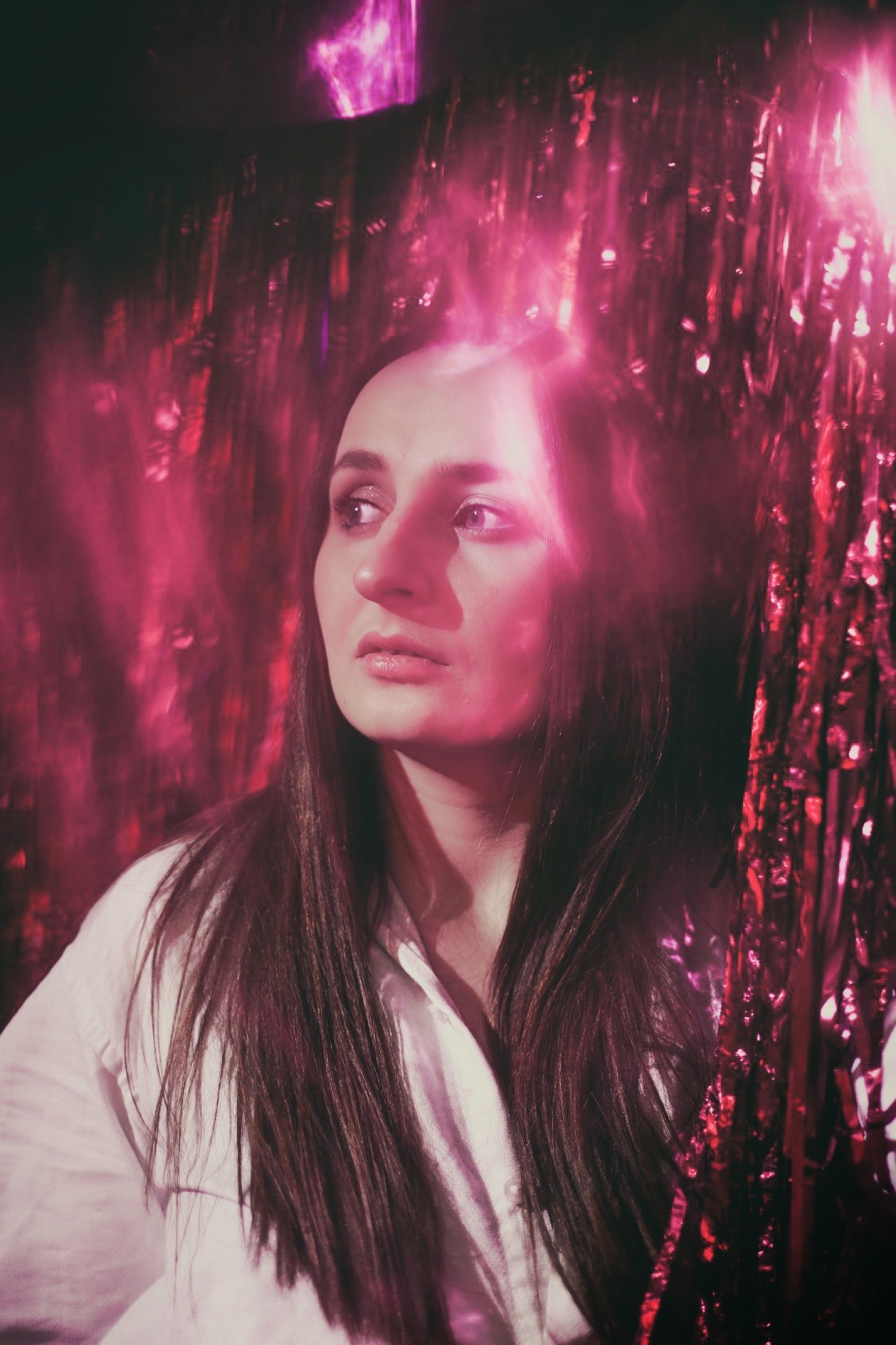 Portret kobiety w białej koszuli, z długimi brązowymi włosami, spoglądającej w bok, z różowym, błyszczącym tłem z frędzli.