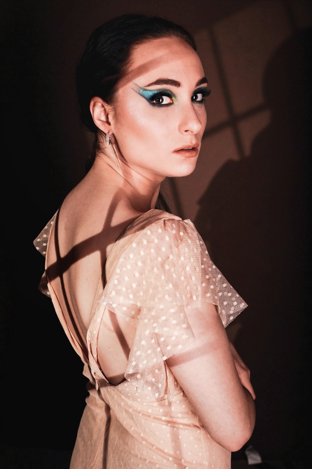 Portret kobiety w sukience w kropki, z intensywnym makijażem oczu w odcieniach niebieskiego i zieleni, z cieniem okien na twarzy i ramieniu, spoglądającej przez ramię w obiektyw.