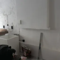 Świeżo otynkowana ściana z widocznymi śladami po instalacjach elektrycznych, biała cegła w tle, przygotowania do dalszych prac wykończeniowych.