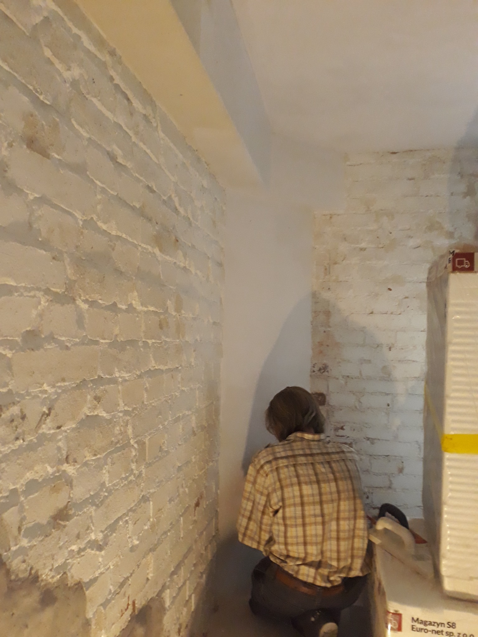 Pracownik w koszuli w kratę szpachluje nierówną, białą ścianę z cegły w pomieszczeniu w trakcie remontu.