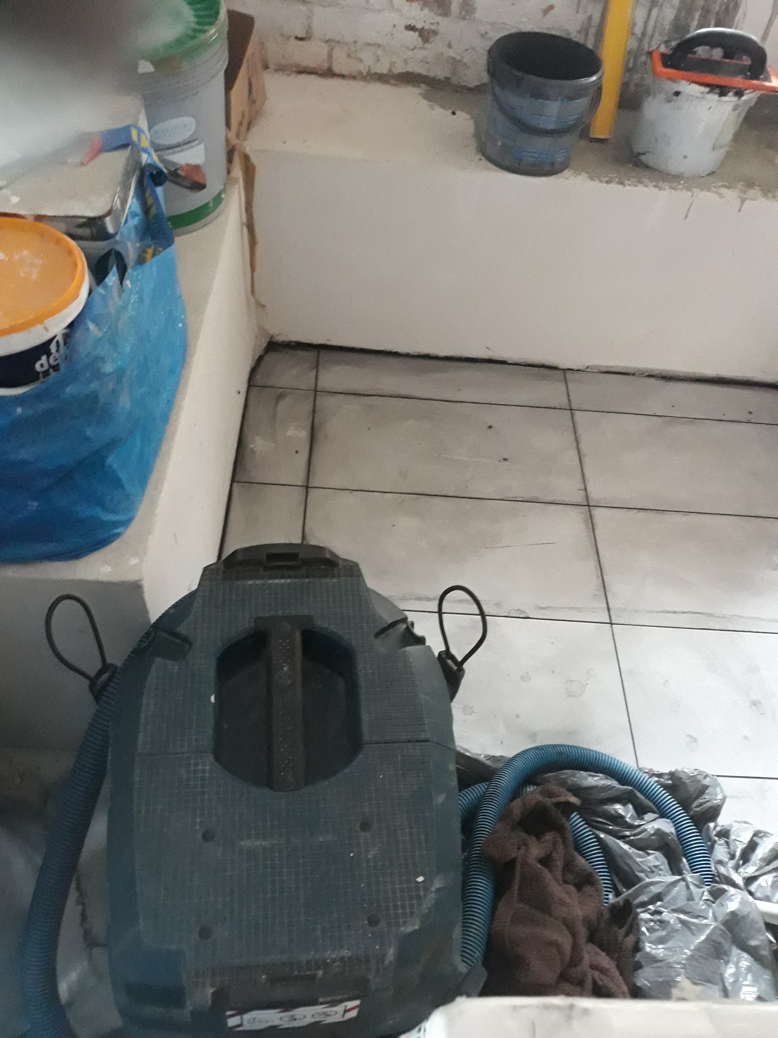 Używany odkurzacz przemysłowy na kafelkach, obok wiadro z farbą, pędzel i niebieski wąż, w tle ściana po remoncie.