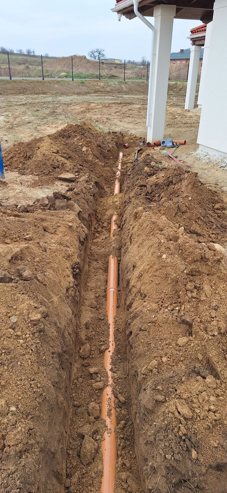 Wykop z ułożonym rurociągiem z tworzywa sztucznego w ziemi, widok z góry. Rury częściowo zasypane, w tle fragment budynku i krajobraz.