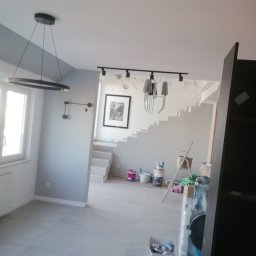 Rem Mod - Wnętrze domu w trakcie remontu, widoczne świeżo pomalowane szare ściany, białe schody, nowoczesne lampy sufitowe i akcesoria malarskie, takie jak pędzle i farby.