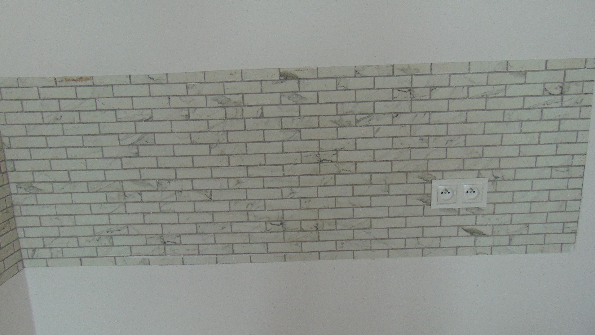 Fragment ściany wykończonej białymi płytkami imitującymi cegłę z szarymi fugami, z zamontowanym podwójnym gniazdkiem elektrycznym.